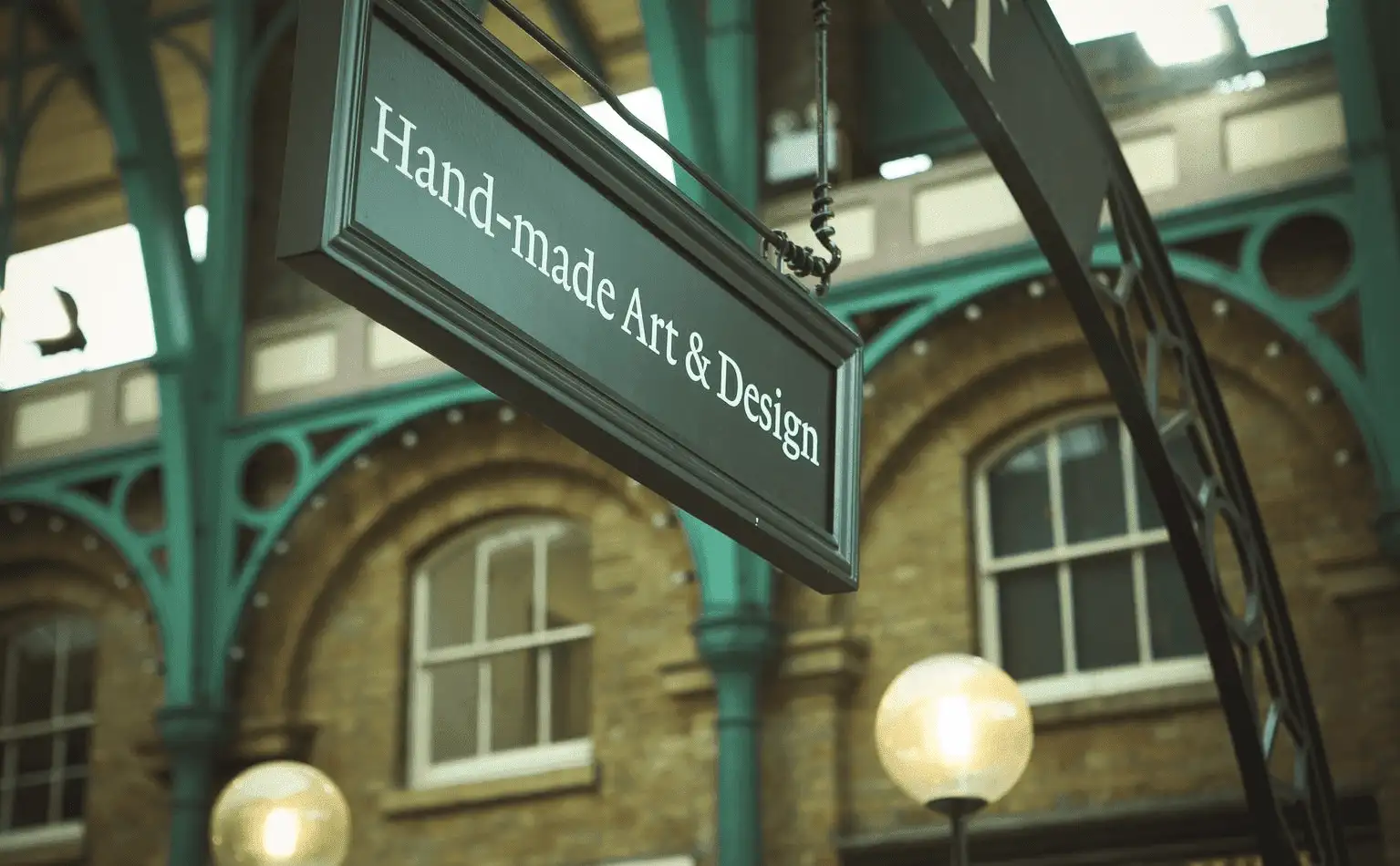 A hanging sign that reads "handmade art & design" in a vintage-style font, set against a backdrop of an arched structure with green metalwork and illuminated globes.