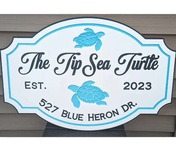 A decorative sign crafted from custom PVC reads The Tip Sea Turtle alongside a turquoise sea turtle graphic. Below, it says Est. 2023 and 527 Blue Heron Dr. The design showcases blue and white hues against a wooden background.