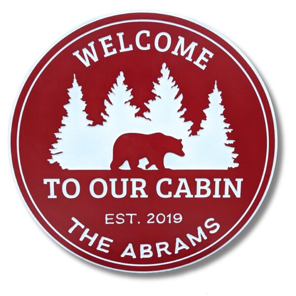 A round red sign with white silhouettes of trees and a bear reads Welcome to Our Cabin and Est. 2019 above The Abrams. This sign, like Personalized Oval Custom Beach House Signs Coastal Decor, merges rustic charm with natures serenity for a warm personal touch.