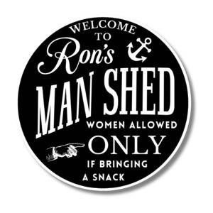 A round black sign with white text reads Welcome to Rons Man Shed and includes an anchor graphic. It playfully evokes coastal decor. Below, Women allowed only if bringing a snack is featured alongside a pointing hand graphic on this Personalized Oval Custom Beach House Sign.