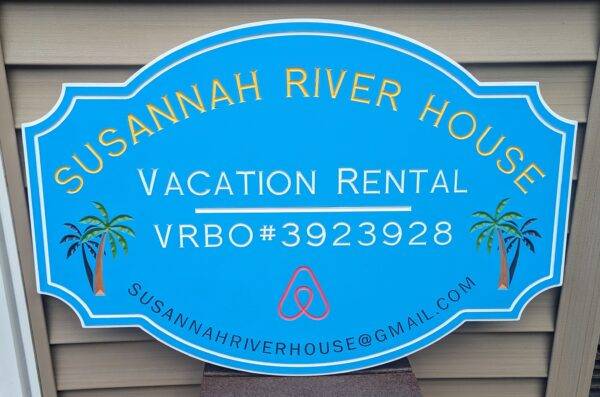 A custom home rental sign for Susannah River House features vibrant yellow and white text on a blue oval background with Vacation Rental VRBO#3923928, an Airbnb logo, palm tree illustrations, and the email address susannahriverhouse@gmail.com.