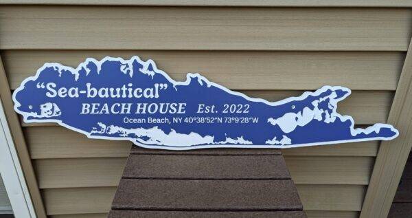 A Long Island-shaped sign in blue and white reads Sea-bautical Beach House Est. 2022 Ocean Beach, NY 40°3852N 73°928W. This Custom Sign for Homes & Rentals is placed on a textured brown surface with a beige background.