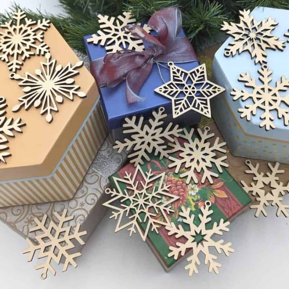 Gift boxes adorned with wooden snowflakes from the Set of 16 Wooden Snowflake Ornaments rest among green pine branches. The hexagonal and rectangular boxes, in blue, gold, and multicolored patterns, showcase a cozy cabin sign theme ideal for festive celebrations.