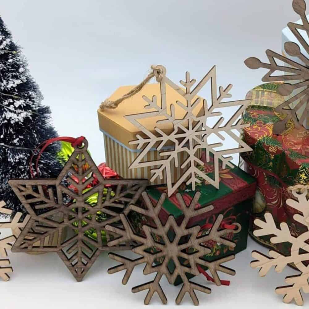 The Set of 6 Extra Large Walnut & Maple Laser-Cut Snowflake Ornaments, arranged with wrapped gifts beside a snow-dusted Christmas tree, evoke cozy cabin charm. Festive papers and a golden-topped gift enhance the warmth of holiday gatherings at your favorite vacation rental.