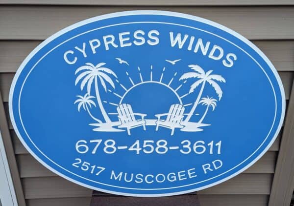 A blue oval PVC sign displays Cypress Winds at the top and 678-458-3611 and 2517 Muscogee Rd at the bottom. The center illustrates two beach chairs, palm trees, and a sun with birds soaring above.