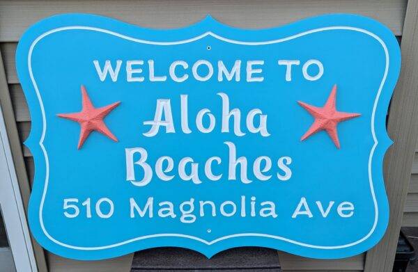 A personalized custom carved 3D PVC sign in blue with white letters says Welcome to Aloha Beaches, 510 Magnolia Ave, and includes two red starfish flanking Aloha Beaches.