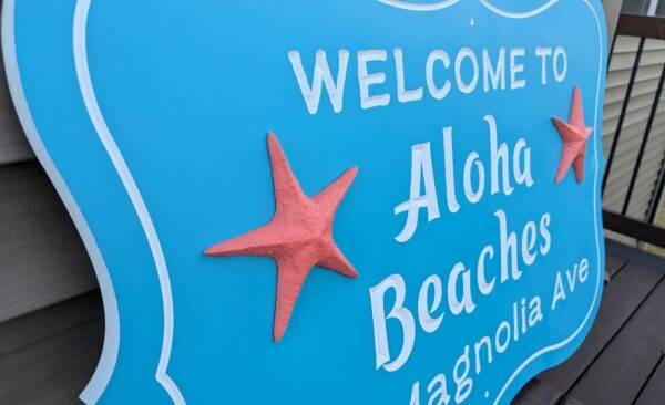 Close-up of a personalized custom carved 3D PVC sign in blue displaying WELCOME TO Aloha Beaches Magnolia Ave. adorned with two red starfish.