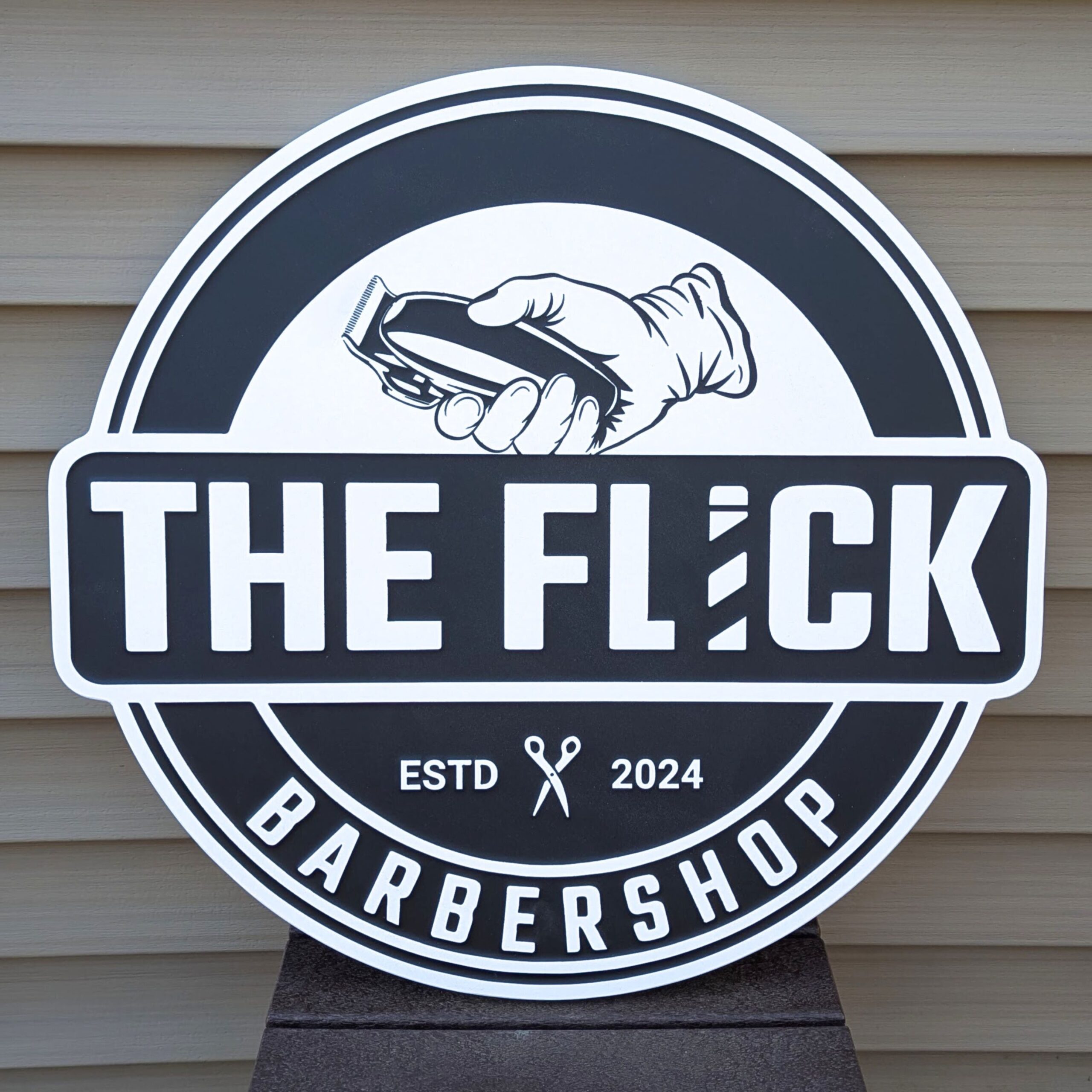 A round barbershop sign reads The Flick Barbershop with an illustration of a hand holding clippers above the text. The sign also says ESTD 2024 with a scissors icon in the center.